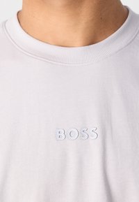 T-shirt gris clair avec un col rond, mettant en avant un logo « BOSS » texturé en matériau blanc légèrement en relief sur la poitrine.