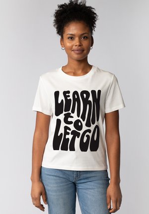 Jonge vrouw met krullend haar, gekleed in een wit T-shirt met vetgedrukte zwarte tekst "LEARN TO LET GO" en een blauwe spijkerbroek, staand tegen een lichte achtergrond.