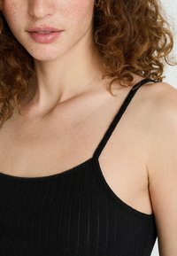 Débardeur noir à côtes avec de fines bretelles et des bords délicatement festonnés. Le tissu a une texture lisse, mettant en valeur une coupe ajustée.