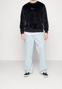 Czarna welwetowa bluza z haftowanym złotym logo, zestawiona z jasnoniebieskimi dżinsami ozdobionymi zakrzywionymi wzorami przeszyć oraz czarnymi trampkami.