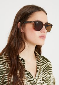 Ochelari de soare tortoise brown cu o formă rotundă, lentile maro închise și logo-ul argintiu Ray-Ban pe brate. Însoțiți de o cămașă cu dungi.