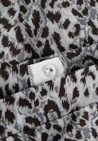 Tissu gris avec un imprimé léopard noir et gris foncé, comportant une étiquette blanche avec un boutonnière cousue et un détail de bouton rond.
