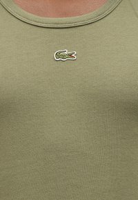 Κοντινό πλάνο υφάσματος με ριγέ υφή σε ανοιχτό ελιά πράσινο, με ραμμένο πράσινο λογότυπο κροκοδείλου και ανοιχτό κόκκινο στόμα στην περιοχή του στήθους.