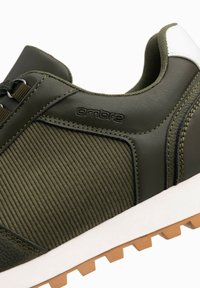 Zielony but sportowy z teksturowaną cholewką, z oliwkowymi paskami, białymi akcentami oraz beżową gumową podeszwą z wyraźnym wzorem bieżnika.