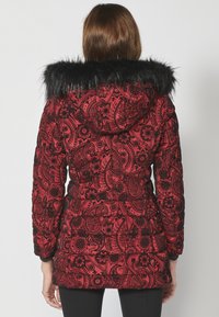 Crvena puffer jakna s crnim cvjetnim paisley uzorkom, s gustom crnom krznom kapuljačom i uskim manžetama. Tekstuirana tkanina s kockastim dizajnom.