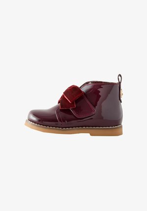 Stivale alla caviglia in pelle verniciata bordeaux lucido con un fiocco in velluto rosso, suola in gomma beige e cucitura decorativa lungo il bordo.