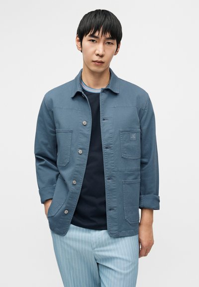 Μπλε βαμβακερό μπουφάν με κουμπιά και δύο μπροστινές τσέπες, φορεμένο πάνω από μια ναυτική μπλούζα, συνδυασμένο με ανοιχτό γαλάζιο παντελόνι με ρίγες. Λείο ύφασμα.