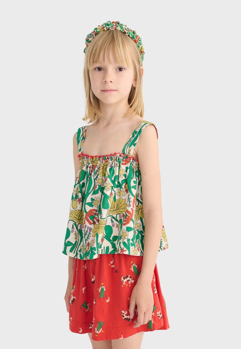 Jeune fille aux cheveux blonds raides portant un haut vert et blanc à fleurs, une jupe rouge avec des imprimés d'animaux de la ferme, et un serre-tête assorti à motif floral.