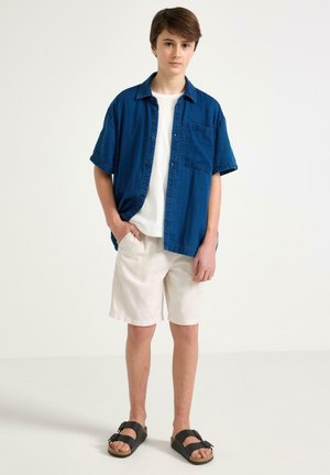 Adolescent debout avec les mains dans les poches, portant une chemise bleue à manches courtes, un T-shirt blanc, un short beige et des sandales noires.