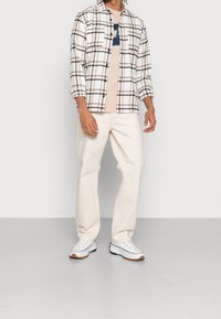 Conjunto de hombre que incluye una camisa de botones blanca a cuadros, una camiseta beige, pantalones crema y zapatillas blancas con detalles marrones.