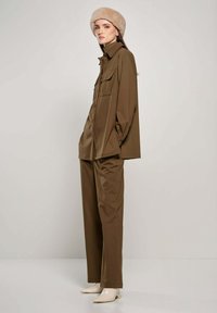 Een bruine blouse met knopen en bijpassende broek, gecombineerd met een beige berenmuts. De stof lijkt soepel en goed passend, wat zorgt voor een samenhangende uitstraling.