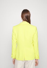 Blazer cintré jaune avec un col texturé et des poignets festonnés. Présente une fente dans le dos pour faciliter les mouvements. Fini en tissu lisse.
