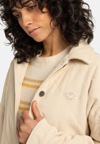 Beige corduroy jacket with a quilted design, featuring a silver snap button and an embroidered logo. Layered over a cream sweater with yellow stripes.