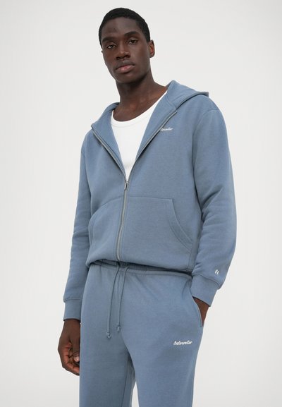 Zilā zip-up kapuce, izgatavota no mīkstas materiāla, ar kenguru kabatu un elastīgiem piedurknēm. Kombinēta ar atbilstošām sporta biksēm, uz kurām ir logo.