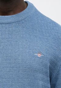 Zils adīts džemperis ar ribotu faktūru, apaļu kakla izgriezumu un nelielu izšūtu sarkani baltu logotipu uz krūtīm.