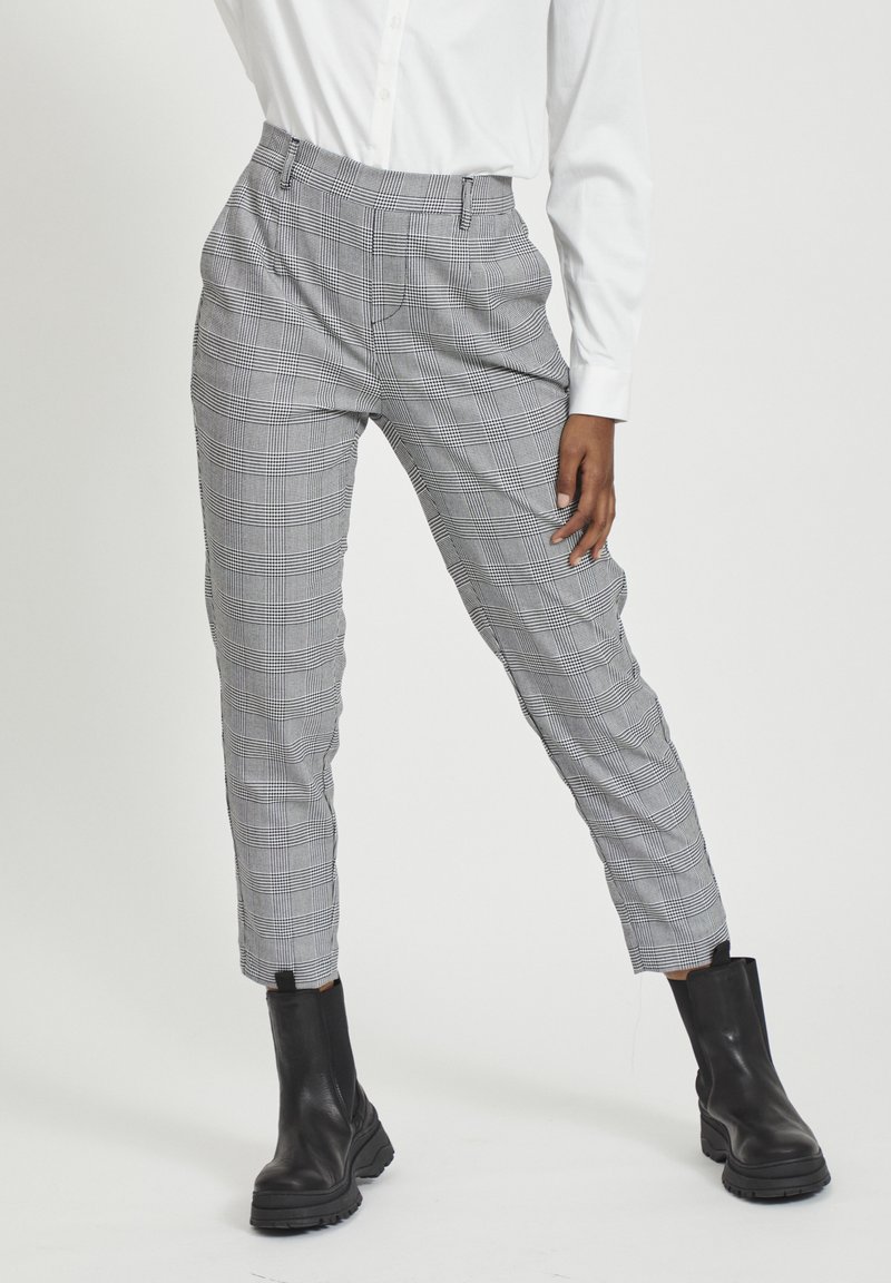 Personne portant un pantalon gris à carreaux fuselé, des bottines noires et une chemise blanche à manches longues, debout devant un fond clair.