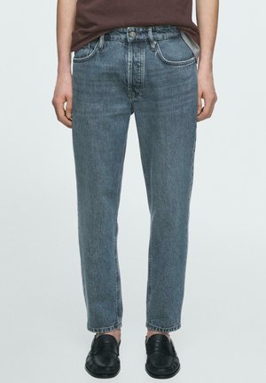 Person iført blå jeans med mellemhøj talje og lige ben, med opkørte buksekanter, kombineret med sorte slip-on loafers, stående mod en ensfarvet baggrund.