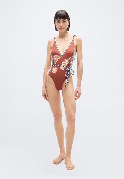 Γυναίκα που στέκεται ξυπόλητη, φορώντας ένα ολόσωμο μαγιό σε σκουριασμένο χρώμα με αφηρημένα λευκά και μπλε μοτίβα και μια ζώνη από δεμένο σχοινί.