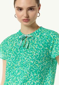 Short-sleeve green blouse with a keyhole opening and turquoise ribbon tie. Features a textured, colorful animal print pattern.