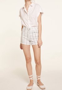 Camisa blanca de manga corta abotonada en el cuello, combinada con pantalones cortos de cuadros de talle alto en blanco y gris. Presenta alpargatas que envuelven el tobillo.