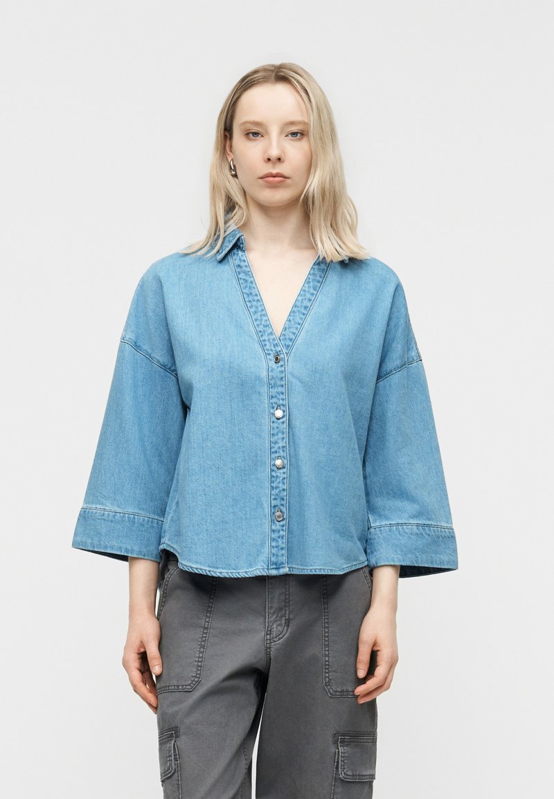 Γυναίκα με ξανθά μαλλιά μέχρι τον ώμο, ντυμένη με ένα χαλαρό ανοιχτό μπλε πουκάμισο από τζιν με κουμπιά και γκρι παντελόνια cargo, στεκόμενη μπροστά σε απλό φόντο.