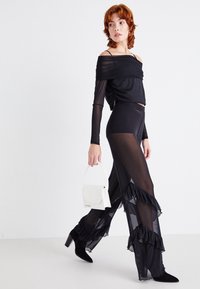 Femme aux cheveux courts roux vêtue d'un haut noir transparent à épaules dénudées et d'un pantalon à volants, tenant un petit sac à main blanc, marchant en studio.