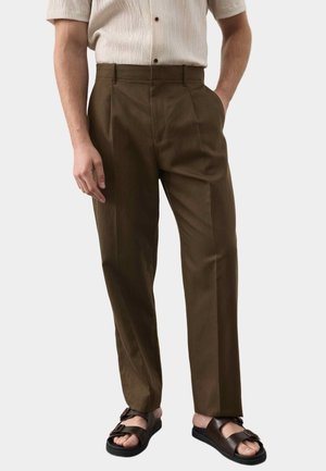 Homme portant un pantalon plissé marron, une chemise crème à manches courtes boutonnée et des sandales noires à boucle, debout les mains dans les poches.