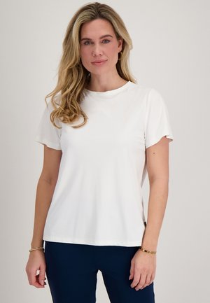 Blonde vrouw met los haar, gekleed in een eenvoudig wit T-shirt met korte mouwen en donkere broek, staand tegen een lichte achtergrond.