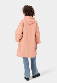 Osoba s kudrnatými vlasy, která má na sobě dlouhý broskvový kabát s kapucí a textem na zádech, světle modré džíny a černé objemné boty, otočená zády.