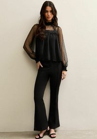 Joven mujer con blusa negra de manga de tul con lunares y pantalones acampanados negros de cintura alta, con tacones abiertos negros, posando frente a una pared beige.