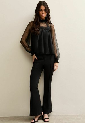 Young woman wearing black sheer polka-dot sleeve blouse and high-waisted black flare pants with black open-toe heels, posing against a beige wall.