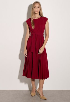 Joven mujer está de pie vistiendo un vestido midi plisado rojo sin mangas con un lazo en la cintura y tacones altos puntiagudos beige, sobre un fondo liso.