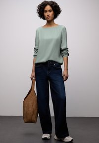Blouse vert clair à manches longues ; jean bleu foncé à jambes larges ; sac fourre-tout en daim beige ; baskets blanches. Tissu lisse, coupe décontractée, look casual.