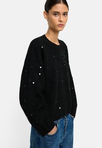 Czarny sweter dzianinowy z cekinami, okrągły dekolt, opuszczone ramiona i luźny krój. Połączony z niebieskimi dżinsami, prezentującymi teksturowany wzór.