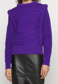 Fioletowy sweter dzianinowy z wysokim dekoltem, teksturowym wzorem w warkocze i ściągaczami przy mankietach. Połączony z czarną skórzaną spódnicą.