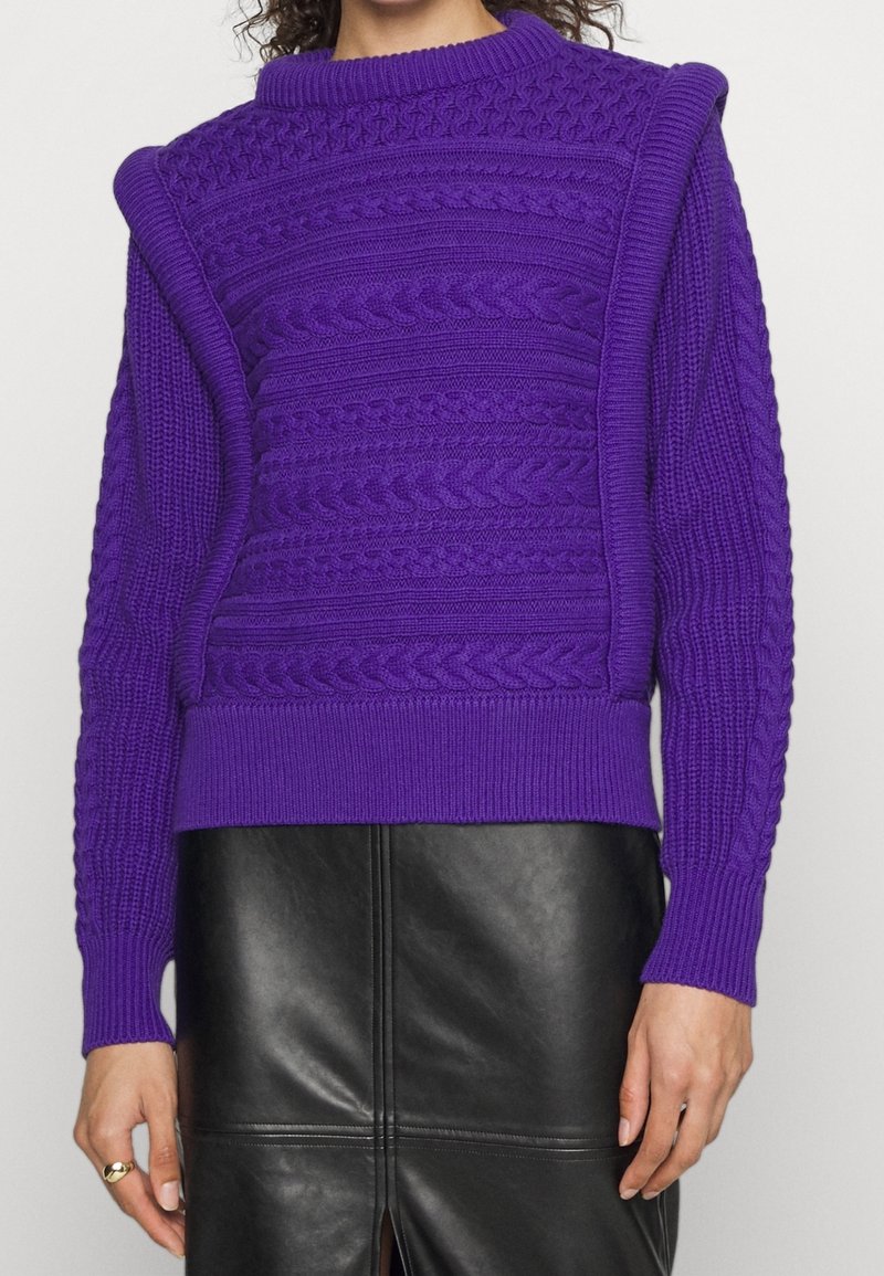 Fioletowy sweter dzianinowy z wysokim dekoltem, teksturowym wzorem w warkocze i ściągaczami przy mankietach. Połączony z czarną skórzaną spódnicą.