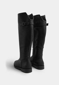 Botas negras hasta la rodilla, hechas de cuero suave con una suela plana. Presentan un cierre lateral de cremallera y un acento de correa en la parte superior.