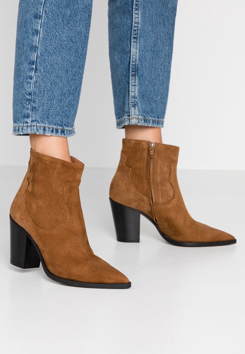 Brune ruskindsankelstøvler med spids tå og skulptureret hæl, med et abstrakt sideløb og lynlåsbaglukning.