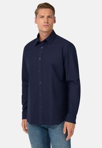 Temno modra srajca z dolgimi rokavi in gumbi, izdelana iz gladkega materiala, z klasičnim ovratnikom in zapenjanjem na sprednji strani, kombinirana z jeans hlačami.