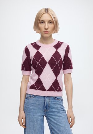 Blonde Frau trägt einen kurzärmeligen pinken und dunkelroten Argyle-Pullover und blaue Jeans und steht vor einem einfarbigen weißen Hintergrund.