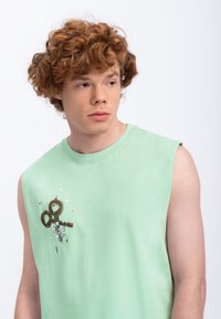 T-shirt sans manches en coton vert clair avec un graphique de ciseaux et des éléments de design abstrait sur le devant. Texture lisse.