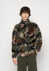 Fleecejakke i camouflage-mønster med nuancer af grøn, brun og sort. Har høj krave og lynlåslukning. Blød tekstur.