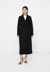 Svart, lang trenchcoat med brede revers, dobbeltspent front, belte i livet, og strukturert stoff. Brukt med sorte høye hæler.