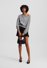 Sudadera gris sin hombros con puños acanalados, combinada con una falda negra. La modelo sostiene un pequeño bolso negro con un logo sutil.