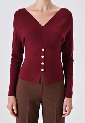 Close-up af en kvinde iført en tætsiddende vinrød V-udskåret sweater med fire knapper foran, kombineret med højtsiddende brune bukser.