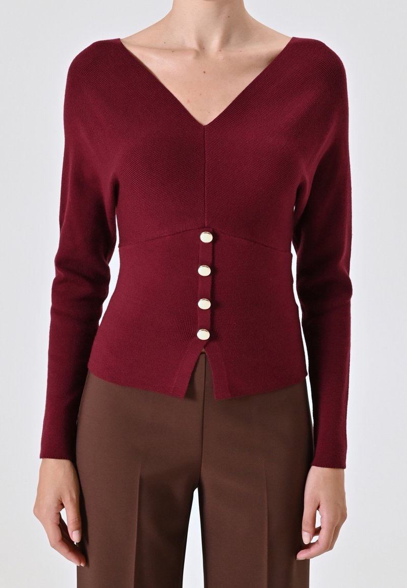 Gros plan d'une femme portant un pull ajusté bordeaux à col en V avec quatre boutons sur le devant, associé à un pantalon marron taille haute.