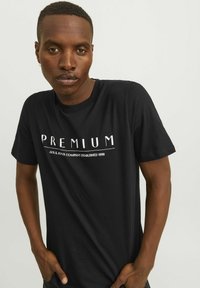 Juodas medvilnės marškinėliai su baltais užrašais "PREMIUM" ir "JACK & JONES COMPANY ESTABLISHED 1990", trumpomis rankovėmis ir klasikinio silueto.