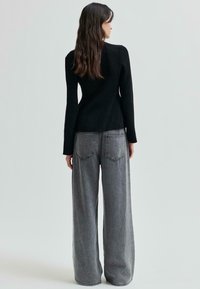 Kvinna med långt mörkt hår står vänd bortåt iförd en åtsittande svart tröja och lösa grå denimjeans mot en enkel bakgrund.