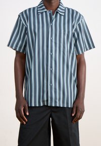 Camisa de botones de manga corta con rayas verticales en azul, verde azulado y blanco, que cuenta con un bolsillo en el pecho y un diseño de cuello tipo camp.