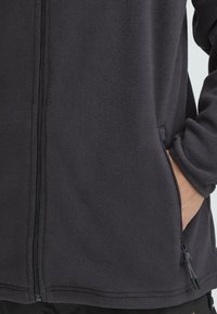 Mustast fliisist jakk, millel on esiosa lukuga sulgur ja küljetaskud. Kangas on pehme ja tekstuuriga, koos elastsete käiste ja lõdva lõikega.